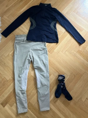 Set med ridbyxor, tröja och jacka - Säljer ett set med beige ridbyxor med förstärkta knän, en svart långärmad träningströja med halv dragkedja och kontrastpaneler samt en lång mörkblå quiltad jacka med huva och röd insida. Perfekt för ridning och utomhusaktiviteter. Använd ett par gånger. 