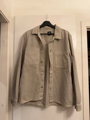 Beige overshirt från Zara - Stilren beige overshirt från Zara i mjuk bomull. Skjortan har två stora bröstfickor, klassisk krage och knäppning framtill. Perfekt att bära öppen över en t-shirt eller stängd för en clean look.