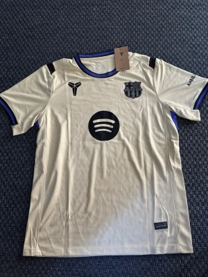 Beige FC Barcelona matchtröja Nike - Snygg beige FC Barcelona matchtröja från Nike med svart klubbmärke, svart sponsorlogga (Spotify) och lila detaljer vid krage och ärmslut. Tillverkad i lätt och ventilerande funktionsmaterial, perfekt för fotboll eller träning. Kortärmad modell med modern passform.