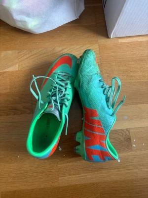 Nike Mercurial gröna fotbollsskor - Snygga Nike Mercurial fotbollsskor i en grön och turkos färg med orange detaljer och logga. Skorna har snörning och dobbar för bra grepp på planen. Tillverkade i syntetmaterial för lätt känsla och bra bollkontroll. Perfekta för dig som vill sticka ut på fotbollsplanen.