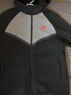 Nike Tech Fleece hoodie svart/grå - Nike Tech Fleece hoodie i svart och grått med hel dragkedja och huva. Ikonisk röd Nike-logga på bröstet och praktiska fickor på sidorna. Tillverkad i mjukt och lätt material som ger en clean och sportig look. Perfekt för dig som gillar streetwear och vill ha något stilrent.