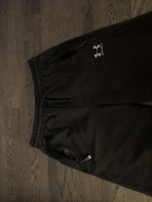  Under Armour shorts - Säljer ett par svarta mjukisbyxor från Under Armour med diskret ribbad struktur. Byxorna har resår i midjan med dragsko, dragkedjeförsedda fickor fram och en bakficka. Perfekta för chill eller träning.