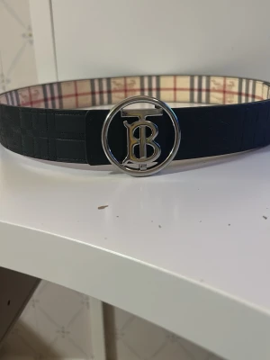 Rutmönstrat bälte med silverdetalj - Snyggt bälte med klassiskt rutmönster i beige, svart och rött samt en rund silverfärgad buckle med bokstaven B. Bältet har en stilren design och passar perfekt till jeans eller byxor för en trendig look.
