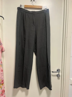 Mörkgrå linnebyxor Vero Moda - Snygga mörkgrå linnebyxor från Vero Moda med raka, vida ben och hög midja. Byxorna har klassisk dragkedja och knapp framtill samt diskreta veck vid linningen. Perfekta för en avslappnad och trendig look.