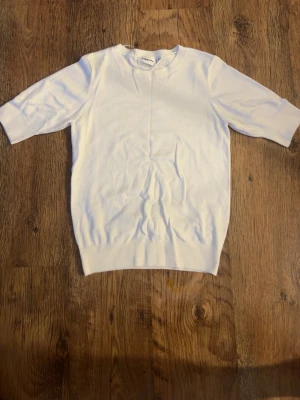 Vit ribbad topp med kort ärm - En stilren vit topp med ribbad nederkant och korta ärmar. Toppen har en enkel, klassisk design och är tillverkad i ett mjukt material som känns skönt mot huden. Perfekt att matcha med jeans eller kjol för en clean look.