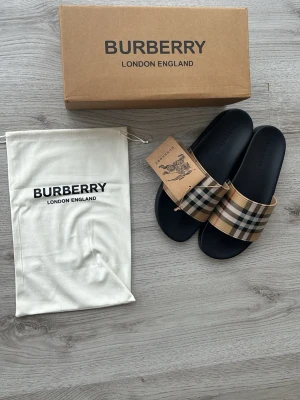 Burberry tofflor  - Säljer ett par snygga Burberry sandaler med klassiskt beige, svart och rött rutigt band över foten. Sulan är svart och har Burberry-loggan präglad. Kommer med originalkartong och dustbag. Perfekta för en trendig och avslappnad stil.