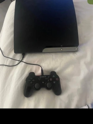 PlayStation 3 Slim med handkontroll - Säljer en svart PlayStation 3 Slim från Sony med tillhörande svart DualShock 3-handkontroll. Konsolen har en stilren, matt finish och rundade kanter. Perfekt för gaming och filmkvällar med vänner.