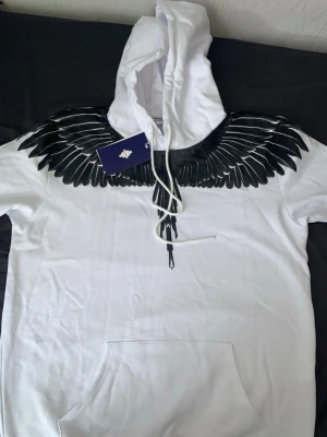 Vit hoodie med svarta vingar från Marcelo Burlon - Säljer en vit hoodie från Marcelo Burlon med ikoniskt svart vingmönster över axlar och bröst. Hoodien har stor huva, dragsko och magficka. Perfekt för dig som vill sticka ut med en unik och trendig design.