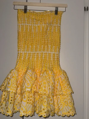 Gul volangklänning Design by Amor - Supersöt gul och vit klänning från Design by Amor med smockad överdel och volangig kjol. Klänningen har broderade detaljer och är perfekt för sommarens alla tillfällen. Tillverkad i ett lätt bomullstyg som känns luftigt och fräscht. Använd 1 gång