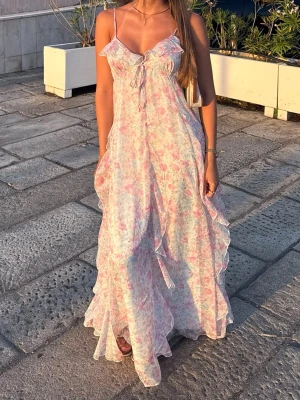 Blommig långklänning med volang - Romantisk långklänning med blommigt mönster i ljusa pastellfärger. Klänningen har tunna axelband, volangdetaljer och knyt framtill. Perfekt för varma sommardagar och har en luftig, flowy passform.