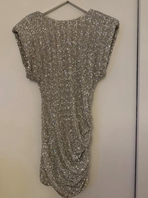 Glittrig silver kortklänning - Säljer en glittrig silverfärgad kortklänning med paljetter och draperad detalj på sidan. Klänningen har en loose passform och är ärmlös, perfekt för dig som vill sticka ut. Supersnygg till fest eller när du vill glänsa lite extra!