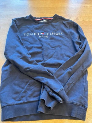 Mörkblå sweatshirt Tommy Hilfiger - Mörkblå sweatshirt från Tommy Hilfiger med broderad logga och text på bröstet. Klassisk rund halsringning med ribbade muddar och nederkant. Snygg detalj med röd och vit rand på insidan av kragen. Perfekt för en avslappnad och stilren look.