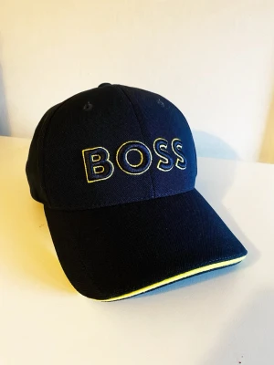 Mörkblå BOSS keps med gul detalj - Snygg mörkblå keps från BOSS med broderad logga i gult framtill. Kepsen har böjd skärm och en gul detalj längs kanten på skärmen. Tillverkad i ett tåligt tyg som ger en sportig och modern look.