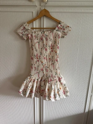Blommig kortklänning med volang - Supersöt kortklänning med blommigt mönster i rosa, grön och gul på vit botten. Klänningen har offshoulder-modell, smockad överdel och volang nedtill. Perfekt för sommarens alla tillfällen!