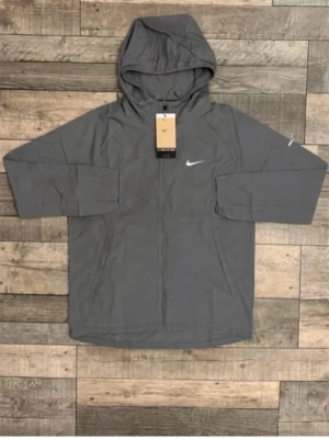 Grå vindjacka med huva från Nike - Snygg grå vindjacka från Nike med dragkedja framtill och justerbar huva. Jackan har två sidofickor och en vit Nike-logga på bröstet. Tillverkad i lätt och vindtätt material, perfekt för blåsiga dagar.