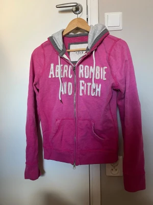 Rosa hoodie från Abercrombie & Fitch - Snygg rosa hoodie med dragkedja från Abercrombie & Fitch. Stor vit logga framtill, grå huva med dragsko och känguruficka. Perfekt för en avslappnad och cool stil. Mjuk bomullskänsla och långa ärmar.
