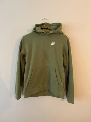 Grön hoodie från Nike med huva - Snygg grön hoodie från Nike med klassisk vit logga på bröstet. Hoodien har en stor känguruficka framtill och en skön huva. Tillverkad i mjukt material och har ribbade muddar vid ärmar och nederkant. Perfekt för en avslappnad och sportig stil.