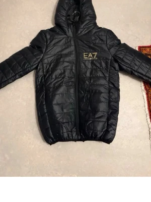 Svart pufferjacka från EA7 Emporio Armani - Snygg svart pufferjacka från EA7 Emporio Armani med huva och guldfärgad logga på bröstet. Jackan har quiltad design, dragkedja framtill och elastiska muddar vid ärmslut och nederkant. Perfekt för kalla dagar och riktigt trendig stil.