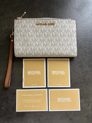 Beige plånbok från Michael Kors - Snygg beige plånbok från Michael Kors med klassiskt MK-mönster och guldfärgad logga framtill. Plånboken har avtagbar handledsrem i brunt läder och dragkedja upptill. Perfekt för att hålla ordning på kort och kontanter. Materialet är tåligt och stilrent.