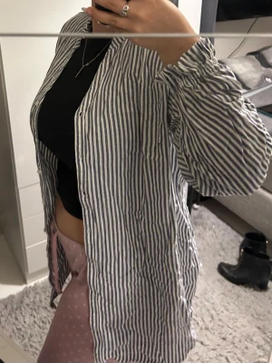 Randig långärmad skjorta i bomull - Säljer en vit och blå randig skjorta i bomull. Skjortan har långa ärmar, knappar framtill och en relaxed, loose passform. Perfekt att slänga över ett linne eller crop top för en chill look.