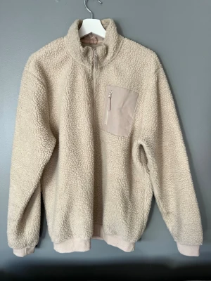 Beige teddytröja från Pier One - Mysig beige teddytröja från Pier One med halv dragkedja och hög krage. Tröjan har en praktisk bröstficka med dragkedja och ribbade muddar vid ärmslut och nederkant. Perfekt för kalla dagar och riktigt skön att bära.