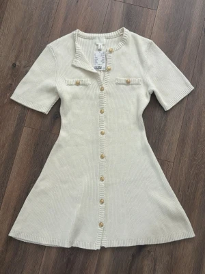 Kort stickad klänning H&M S - Kort stickad klänning från H&M med korta ärmar, dekorativa knappar framtill och två fejkfickor på bröstet. Klänningen har en lätt utsvängd passform och ribbade detaljer vid krage och knappslå. Perfekt för en trendig och avslappnad look.
