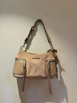 Beige axelremsväska Steve Madden - Snygg beige handväska från Steve Madden med justerbar axelrem och två framfickor med dragkedja och långa läderdetaljer. Väskan har en mjuk, rundad form och är tillverkad i ett slätt material med metalliska detaljer och logga framtill.
