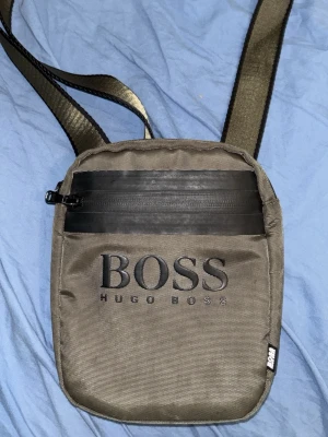 Grön Axelväska från Hugo Boss i svart - Snygg grön axelväska från Hugo Boss med justerbar rem och dragkedja framtill. Väskan har tydlig BOSS-logga på framsidan och är tillverkad i slitstarkt tygmaterial. Perfekt för att bära det viktigaste och ger en clean streetwear-look.