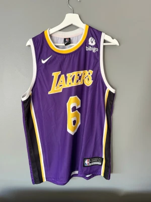 Lakers basketlinne Nike XL - Snyggt basketlinne från Nike med Los Angeles Lakers tryck och nummer 6. Linnet är lila med gula och vita detaljer, samt svarta sidopaneler. Tillverkat i lätt och ventilerande material, perfekt för basket eller streetwear.