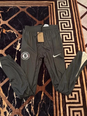 Grå Chelsea träningsbyxor Nike - Snygga grå träningsbyxor från Nike med Chelsea FC-logga på vänster ben och vit Nike Swoosh på höger ben. Byxorna har smal passform, resår i midjan och dragkedjor vid bensluten. Perfekta för fotbollsträning eller chill.