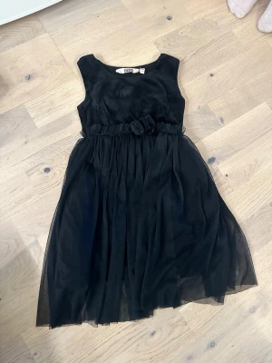 Svart ärmlös tyllklänning med rosett - Snygg svart klänning med tyllkjol och markerad midja med en söt rosettdetalj. Klänningen är ärmlös och har en rund halsringning. Perfekt för dig som vill ha en klassisk look med lite extra volym i kjolen.Svart ärmlös tyllklänning med rosett 