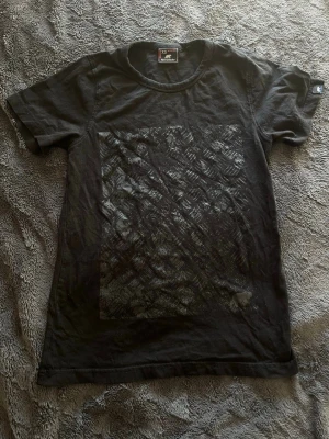 Svart t-shirt från Björnkläder XS - Svart t-shirt från Björnkläder i storlek XS. T-shirten har ett diskret rutmönster framtill och klassisk rund hals. Perfekt för en avslappnad och stilren look.