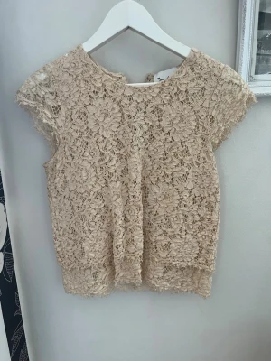 Beige spetsblus med blommönster - Supersöt beige blus i spets med blommigt mönster och korta ärmar. Perfekt att styla med jeans eller kjol för en trendig look. Blusen har rund halsringning och är genombruten i materialet vilket ger en luftig känsla.