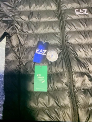 Svart dunjacka från EA7 Emporio Armani - Snygg svart dunjacka från EA7 Emporio Armani med huva och dragkedja framtill. Jackan har ett quiltat mönster och logga på bröstet. Perfekt för vår&sommsr och ger en sportig look. Materialet är lätt och smidigt, vilket gör den enkel att bära.