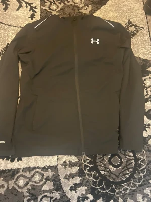 Under Armour vindjacka - Svart träningsjacka från Under Armour med dragkedja framtill och två sidofickor. Jackan har långa ärmar, diskreta reflexdetaljer på axlarna och logga på bröstet. Tillverkad i ett tekniskt material som passar perfekt för träning utomhus.
