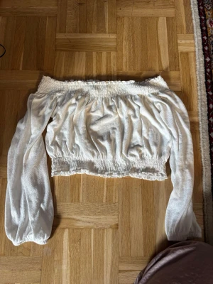 Vit offshoulder blus med långa ärmar - Trendig vit blus med offshoulder-design och långa, luftiga ärmar. Smockad upptill och nertill för en snygg passform. Perfekt för dig som gillar en romantisk och avslappnad stil.