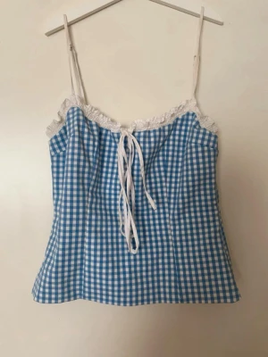 Blåvit rutig korsett med snörning Zara - Supersöt korsett topp i blåvitt rutigt mönster med smala axelband och vit spetskant upptill. Snörning framtill ger en extra detalj och toppen har en figurnära passform. Perfekt för dig som vill ha en trendig och feminin look.