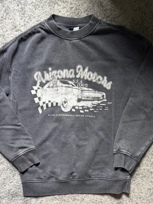 Grå sweatshirt med Arizona Motors-tryck - Cool grå sweatshirt med vintageinspirerat Arizona Motors-tryck framtill. Tröjan har rund hals, ribbade muddar och avslappnad passform. Perfekt för dig som gillar retrostil och motorsport. Materialet känns mjukt och bekvämt mot huden.