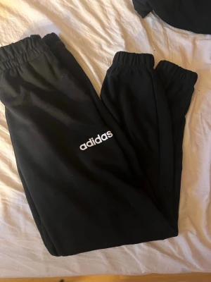 Svarta mjukisbyxor från Adidas - Svarta mjukisbyxor från Adidas med vit logga på benet. Byxorna har resår i midjan och vid bensluten för en skön och avslappnad passform. Perfekta för chill eller träning. Tillverkade i mjukt material som känns bekvämt hela dagen.