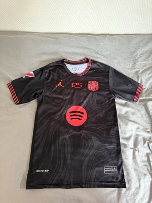 Svart FC Barcelona matchtröja - Säljer en svart FC Barcelona matchtröja med röda detaljer och tryck. Tröjan har korta ärmar, rund hals och är tillverkad i Dri-FIT material. På bröstet finns Spotify-logga, Jordan-logga och klubbmärke. På ryggen står Lamine Yamal och nummer 10 i rött.