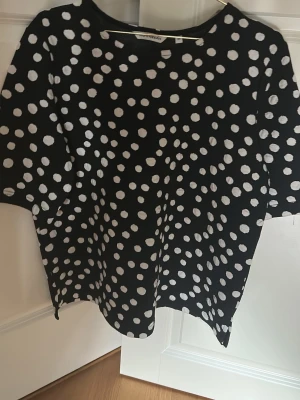 Svart prickig topp från KappAhl - Snygg svart topp från KappAhl med vita prickar. Modellen har rund halsringning och korta ärmar. Perfekt för dig som gillar ett lekfullt mönster och vill ha en bekväm och stilren topp i garderoben.