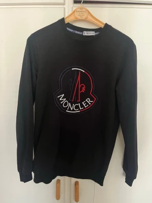 Svart Moncler sweatshirt med logga - Svart sweatshirt från Moncler med stor broderad logga i vitt och rött framtill. Tröjan har rund halsringning och långa ärmar. Perfekt för dig som gillar stilrena och exklusiva plagg med tydlig branding.