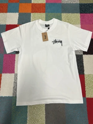 Vit Stüssy t-shirt med tryck - Säljer en vit t-shirt från Stüssy med svart logga på bröstet och stort tryck med rosa tärningar och logga på ryggen. Klassisk passform och korta ärmar. Perfekt för dig som gillar streetwear och vill sticka ut lite extra.