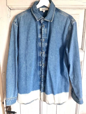 Blå jeansskjorta från COS - Säljer en snygg blå jeansskjorta från COS med vit dip dye-detalj längst ner. Skjortan har klassisk krage, långa ärmar och knappar framtill. Perfekt för dig som gillar en avslappnad men trendig look.