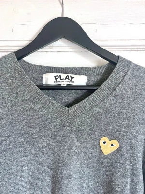 Grå stickad tröja Comme des Garçons Play - Grå stickad tröja från Comme des Garçons Play med V-ringning och lång ärm. Tröjan har ett gult hjärta med ögon broderat på bröstet. Klassisk och enkel design med ribbade muddar och nederkant. Perfekt för dig som gillar streetwear och stilrena plagg.