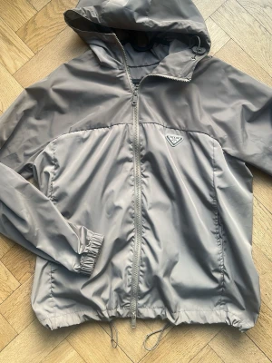Prada Grå/Grön Wind Jacket - Snygg grå vindjacka från Prada med dragkedja framtill, justerbar nederkant och elastiska muddar vid ärmarna. Jackan har en klassisk huva och den ikoniska Prada-loggan på bröstet. Tillverkad i ett lätt och vattenavvisande material som passar perfekt till alla tillfällen. 