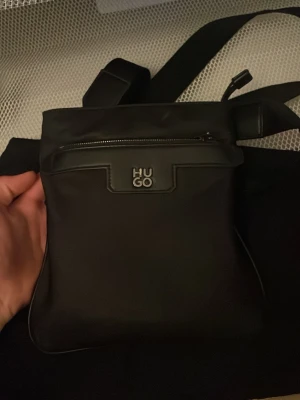 Svart axelväska från HUGO - Snygg svart axelväska från HUGO med justerbar rem och dragkedja framtill. Väskan har en stilren design med metallogo på framsidan och är tillverkad i ett slätt material som känns modernt och enkelt att matcha.