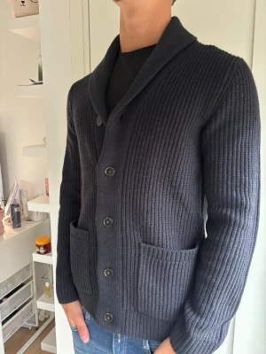 Mörkgrå ribbstickad kofta - Snygg mörkgrå ribbstickad kofta med sjalkrage och knäppning framtill. Två stora fickor framtill och långa ärmar. Perfekt till jeans för en chill och stilren look.