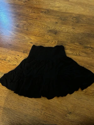 Svart kortkjol med volang - Säljer en svart kortkjol med bred smock i midjan och volangig nederkant. Kjolen har en luftig och flowy känsla, perfekt för dig som gillar en enkel men snygg stil.