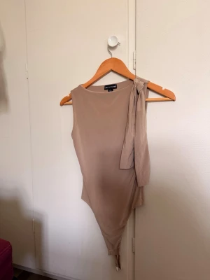 Beige body med knytband - Snygg beige body från PrettyLittleThing med ett brett knytband på ena axeln. Tillverkad i ett mjukt, stretchigt material som sitter skönt mot kroppen. Sitter som ”hud” (tajt) på kroppen. Aldrig kommit till andvändning